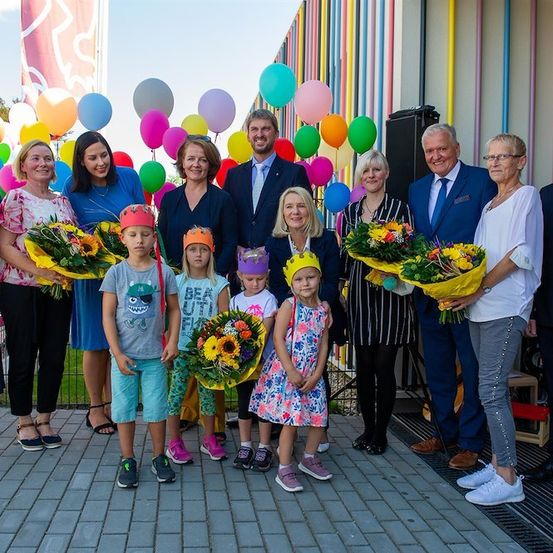 Eine Gruppe von Erwachsenen und Kindern steht vor einer bunten Wand mit Luftballons, hält Blumen und trägt Partyhüte.