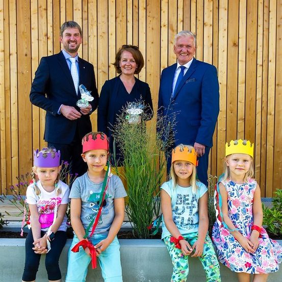 Eine Gruppe von Menschen, einschließlich Erwachsener und Kindern, steht vor einer Holzwand und lächelt für ein Foto. Die Kinder tragen Kronen und halten Blumen.