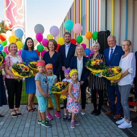 Eine Gruppe von Erwachsenen und Kindern steht vor einem farbenfrohen Gebäude und hält Blumensträuße. Die Erwachsenen sind formell gekleidet, und die Kinder tragen Partyhüte. Dahinter sind bunte Luftballons an einem Zaun befestigt.