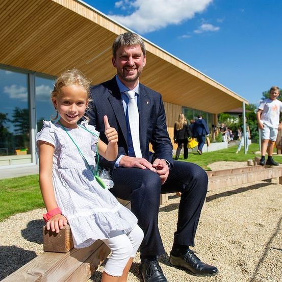 Ein Mann in Anzug und Krawatte sitzt auf einer Bank mit einem kleinen Mädchen, beide lächeln. Hinter ihnen steht ein modernes Gebäude mit Glasfenstern und einem Rasenbereich. Im Hintergrund stehen und gehen einige Personen.