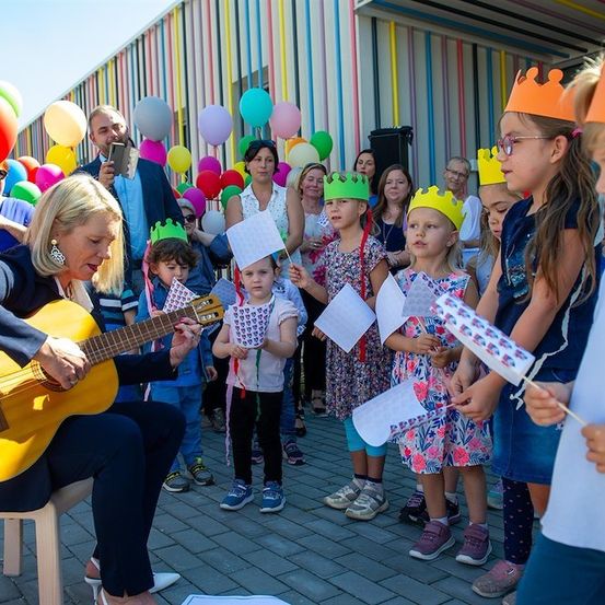 Eine Frau spielt Gitarre für eine Gruppe von Kindern mit Kronen, die Flaggen halten, umgeben von Luftballons und Erwachsenen.