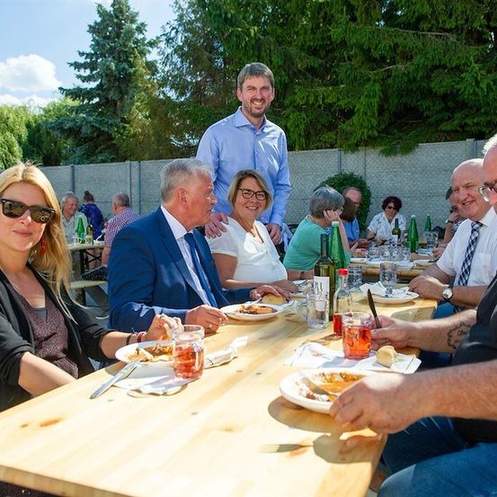 Eine Gruppe von Menschen sitzt um einen Tisch in einem Outdoor-Essbereich. Ein Mann steht hinter ihnen und lächelt. Der Tisch ist mit Tellern, Gläsern und Flaschen gedeckt. Die Umgebung ist im Freien, mit einem Zaun und Bäumen im Hintergrund.