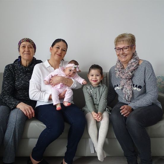 Vier Frauen und ein Baby sitzen auf einer Couch und lächeln für ein Foto. Das Baby sitzt in der Mitte, gehalten von einer der Frauen.