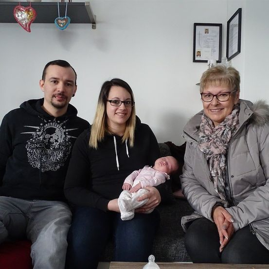 Eine Familie mit drei Erwachsenen und einem Baby posiert für ein Foto auf einem Sofa. Der Mann trägt eine schwarze Kapuzenjacke, die Frau hält das Baby und die ältere Frau trägt eine Brille und einen Mantel.