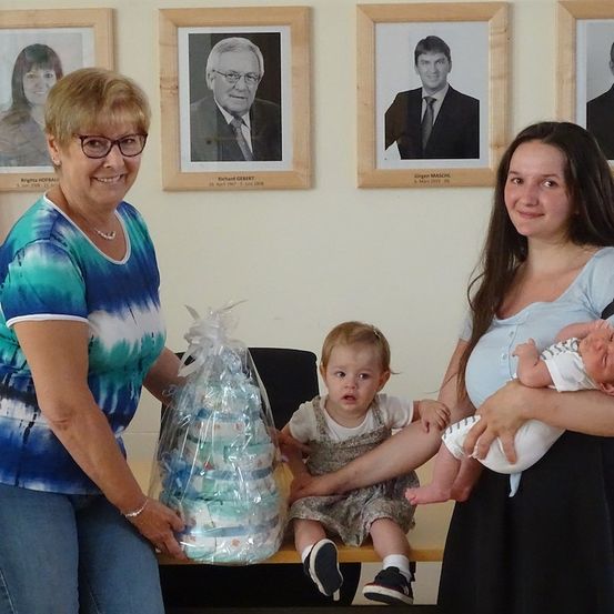 Zwei Frauen halten ein Baby und einen Geschenkkorb. Eine Frau hält ein Baby und die andere einen Korb mit Windeln. Hinter ihnen hängen drei gerahmte Porträts an der Wand.