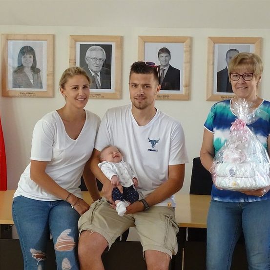 Eine Familie aus drei Personen, darunter ein Mann, der ein Baby hält, steht vor einer Wand mit gerahmten Porträts. Die Frau links trägt zerrissene Jeans, der Mann hält ein Baby und die Frau rechts hält ein eingewickeltes Geschenk.