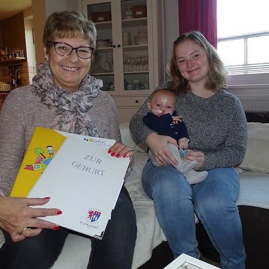 Eine Frau mit einer Urkunde und einem Buch sitzt auf einem Sofa mit einem Baby auf dem Schoß. Eine andere Frau mit Brille sitzt neben ihr.