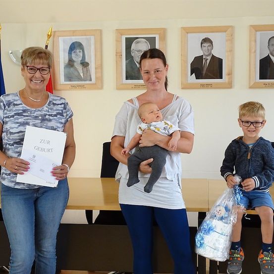 Drei Personen stehen vor einer Wand mit gerahmten Porträts. Eine Frau hält ein Zertifikat, eine andere Frau ein Baby und ein Junge eine Tasche.