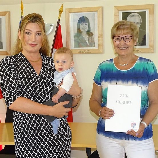 Eine Frau, die ein Baby hält, steht neben einer anderen Frau, die ein Zertifikat mit der Aufschrift 'Zur Geburt' hält. Sie lächeln beide und stehen vor einer Wand mit gerahmten Fotos und Flaggen.