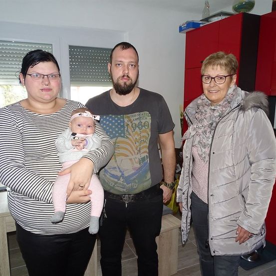 Drei Personen stehen in einem Raum, eine hält ein Baby, mit einer Frau in einem gestreiften Shirt, einem Mann in einem grauen T-Shirt und einer älteren Frau in einem grauen Mantel.