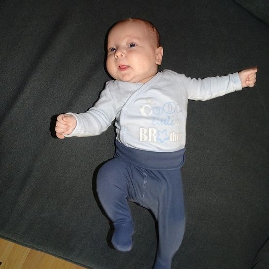 Ein Baby, das ein weißes Langarmshirt mit der Aufschrift 'Cool little BRother' trägt, liegt auf einer dunklen Oberfläche und schaut nach oben.