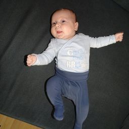 Ein Baby, das ein weißes Langarmshirt mit der Aufschrift 'Cool little BRother' trägt, liegt auf einer dunklen Oberfläche und schaut nach oben.