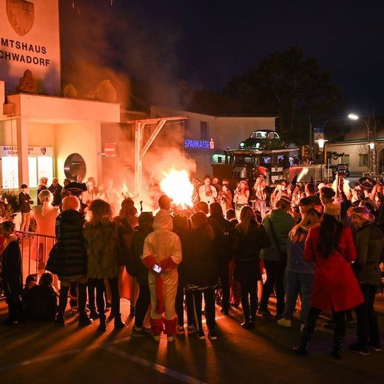 Eine Menschenmenge versammelt sich bei Nacht um ein Lagerfeuer, mit einem Gebäude namens 'Amtshaus Schwadorf' im Hintergrund. Einige Personen tragen Kostüme, und ein Feuerwehrauto ist in der Nähe geparkt.