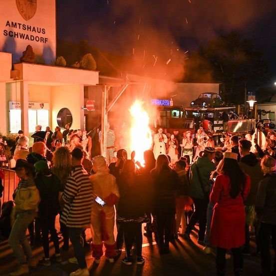 Eine Menschenmenge in Kostümen versammelt sich nachts um ein Lagerfeuer vor einem Gebäude mit dem Schild 'Amtshaus Schwadorf'.