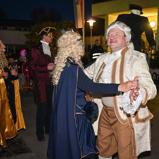 Ein Mann und eine Frau in historischen Kostümen tanzen bei Nacht auf der Straße, umgeben von anderen kostümierten Personen.