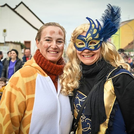 Zwei Frauen in Kostümen, eine in einem Giraffen-Overall und die andere mit einer blauen Federmaske, lächeln für ein Foto bei einer Veranstaltung im Freien.