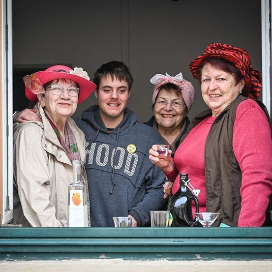Vier Personen stehen lächelnd in einem Fenster. Zwei Frauen tragen Hüte, eine mit einer Blume. Ein Mann in einer Kapuzenjacke hält ein Getränk. Eine Flasche und Gläser stehen auf der Fensterbank.