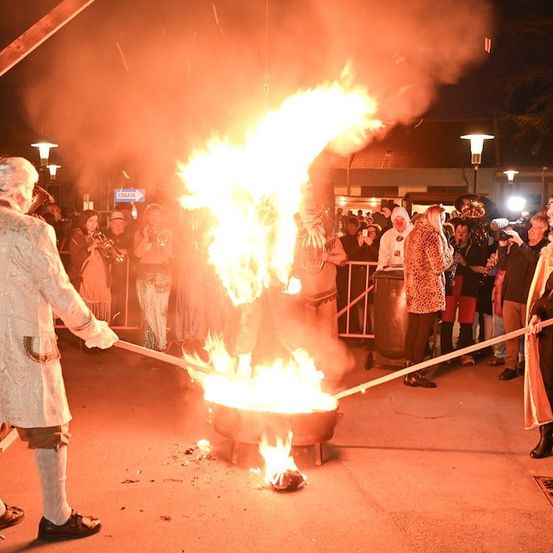 Ein Mann und eine Frau in Kostümen rühren mit langen Stäben in einem großen Topf Feuer, umgeben von einer Menge Zuschauer.