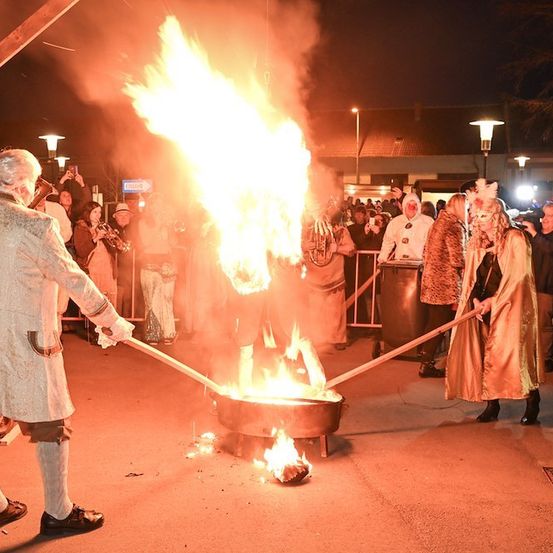 Ein Mann und eine Frau in Kostümen rösten Marshmallows über einem Feuer, während eine Menge zuschaut.