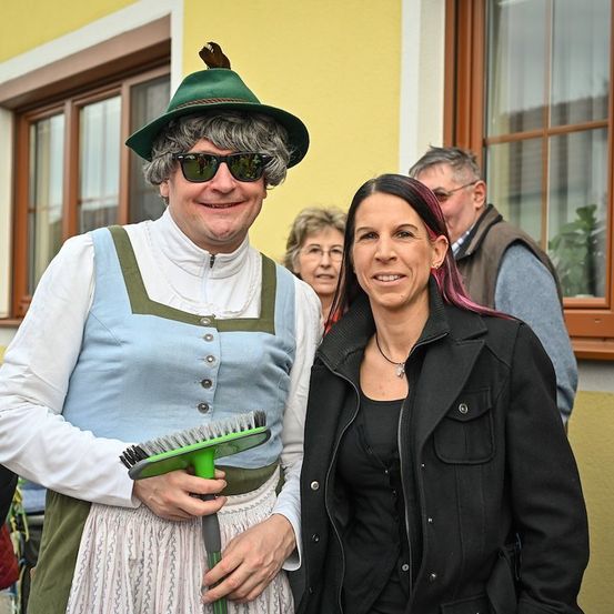 Ein Mann in traditioneller bayerischer Kleidung hält eine Besen und steht neben einer lächelnden Frau in einer schwarzen Jacke. Dahinter stehen ein Mann und eine Frau in der Nähe eines Fensters.