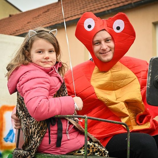 Ein kleines Mädchen in einer rosa Jacke und einem Leoparden-Kleid hält einen Luftballon, während es auf dem Schoß eines Mannes sitzt, der in einem roten und gelben Schlangenkostüm gekleidet ist.