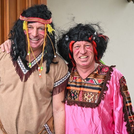 Zwei Männer in farbenfrohen Kostümen, einer in einer braunen Tunika mit Kopfschmuck und der andere in einem rosa Kleid mit rotem und schwarzem Kopfschmuck, lächeln und posieren für ein Foto.