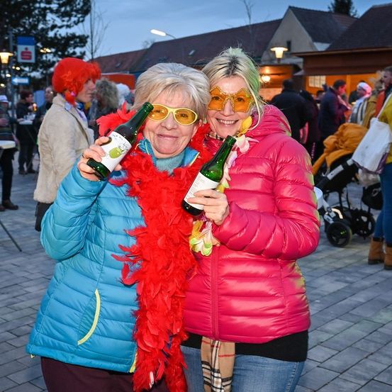 Zwei Frauen in Wintermänteln, eine hält eine grüne Flasche und die andere eine pinke Flasche, lächeln und posieren für ein Foto in einer festlichen Umgebung im Freien.