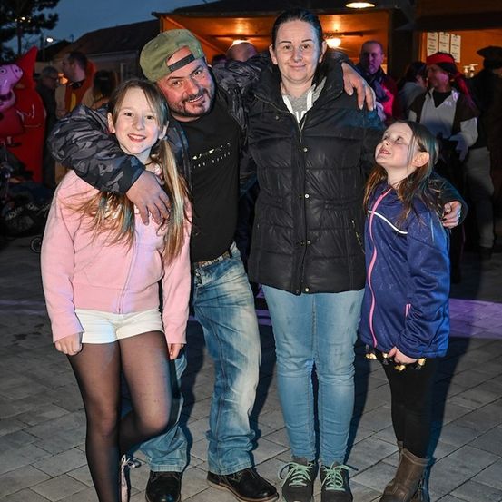 Eine Familie aus vier Personen, bestehend aus zwei Mädchen und zwei Erwachsenen, posiert für ein Foto im Freien bei Nacht. Der Mann trägt eine Camouflage-Jacke und eine Mütze, während die Frau eine schwarze Jacke trägt. Die beiden Mädchen tragen rosa und blaue Jacken.