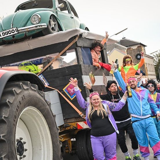 Eine Gruppe von Menschen in bunten 80er-Jahre-Outfits posiert für ein Foto vor einem Traktor mit einem grünen Auto obenauf.