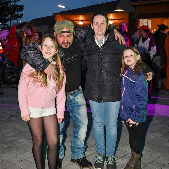 Eine vierköpfige Familie steht bei Nacht und lächelt für die Kamera. Der Mann trägt eine Mütze und hat seinen Arm um die Frau, die eine schwarze Jacke trägt. Die beiden Mädchen, eines in Pink und eines in Blau, stehen neben ihnen. Im Hintergrund sind Menschen und ein Gebäude mit Lichtern zu sehen.