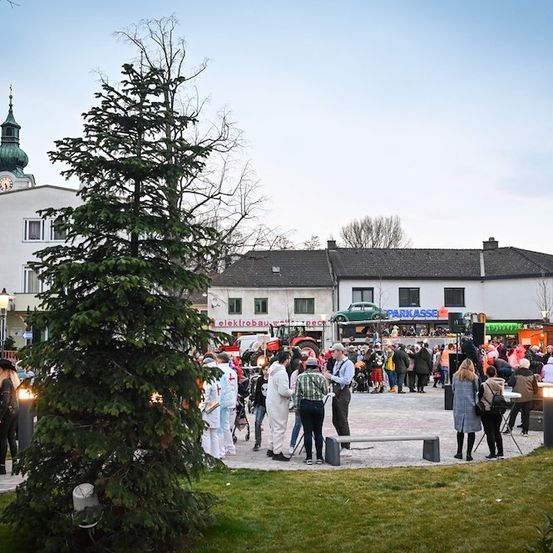 Eine große Menschenmenge in Kostümen versammelt sich auf einem Stadtplatz, mit einem großen immergrünen Baum und einem Uhrturm im Hintergrund.