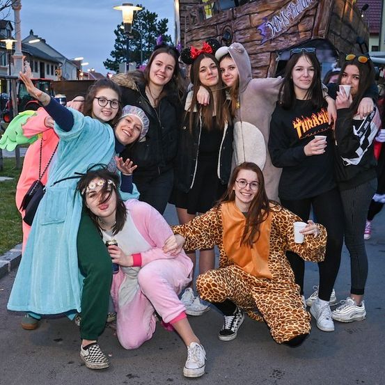 Eine Gruppe junger Frauen in verschiedenen Kostümen posiert für ein Foto auf einer Straße bei Nacht. Einige tragen Tierkostüme, andere Hoodies und Brillen. Sie lächeln und halten Tassen. Hinter ihnen steht eine große, dekorierte Struktur.