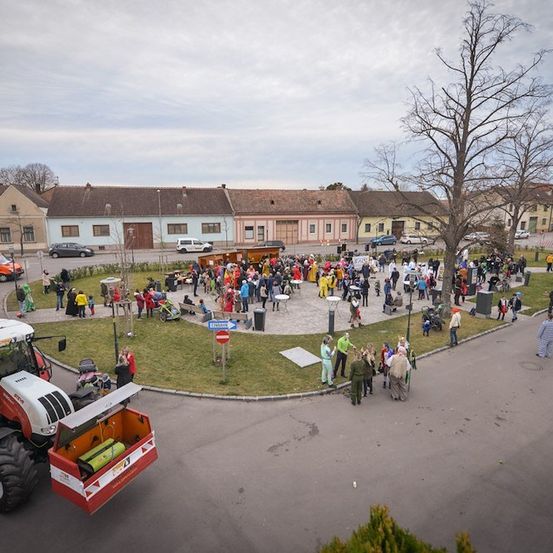 Eine Vogelperspektive eines Stadtplatzes mit einer großen Menschenmenge, einige in Kostümen gekleidet, versammelt um einen zentralen grünen Bereich mit einem Traktor links. Gebäude säumen den Rand, und ein bewölkter Himmel ist über ihnen.