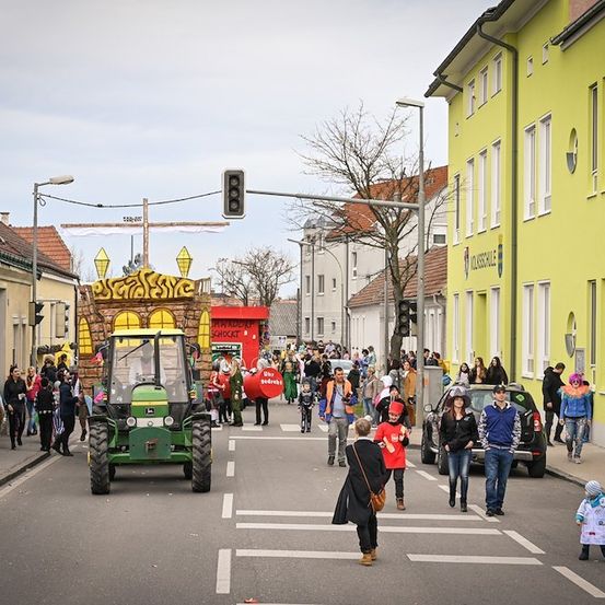 Ein Straßenumzug mit einem geschmückten Traktor an der Spitze, Menschen in Kostümen auf der Straße und Gehwegen und Gebäuden auf beiden Seiten.