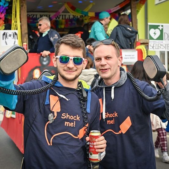 Zwei Männer in blauen T-Shirts mit dem Text 'Shock me!' halten ein schnurgebundenes Telefon in einer Parade. Dahinter sind ein bunter Wagen und Menschen zu sehen.