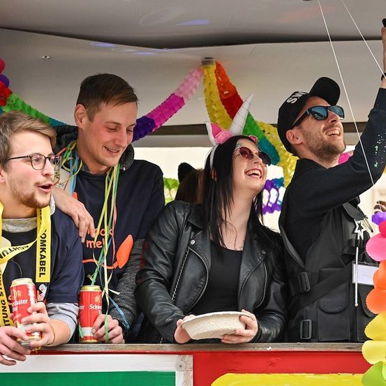 Eine Gruppe junger Erwachsener auf einem Festival, einer macht ein Selfie, während andere Getränke und einen Teller halten. Sie sind von bunten Dekorationen umgeben.