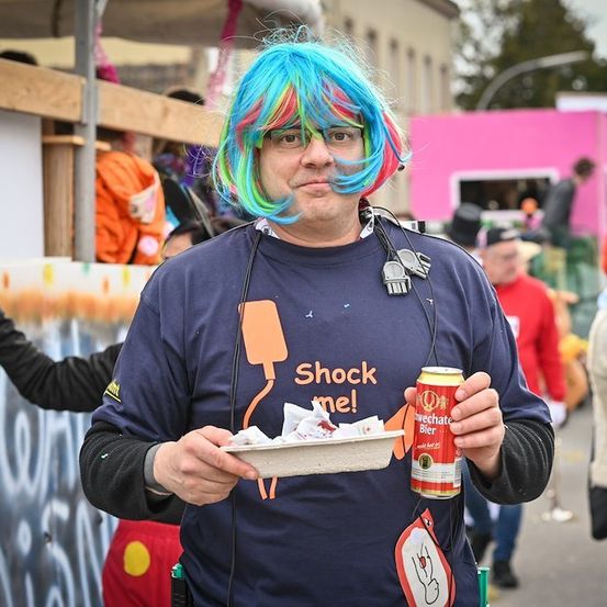 Ein Mann mit blauen und rosafarbenen Locken, der ein blaues Shirt mit dem Aufdruck 'Shock me!' trägt, eine Bierdose und einen Teller mit Essen in der Hand hält.