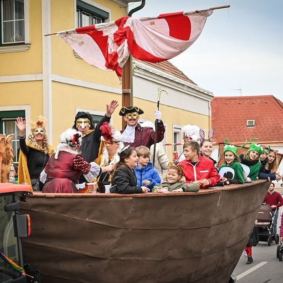 Eine Gruppe von Menschen in Kostümen fährt auf einem Umzugswagen und winkt und lächelt. Der Wagen sieht aus wie ein Schiff mit einer Flagge auf einem Mast. Gebäude und Zuschauer säumen die Straße.