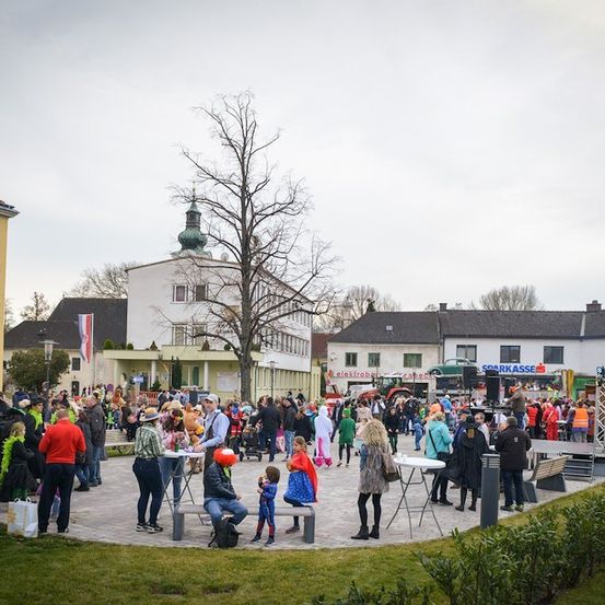 Eine große Menschenmenge in Kostümen und Hüten versammelt sich auf einem Stadtplatz, mit Gebäuden und einem Baum im Hintergrund.
