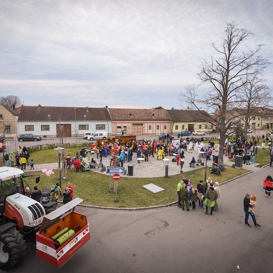 Luftaufnahme eines lebhaften Stadtplatzes mit einem Traktor, einer Menschenmenge und verschiedenen Gebäuden im Hintergrund.