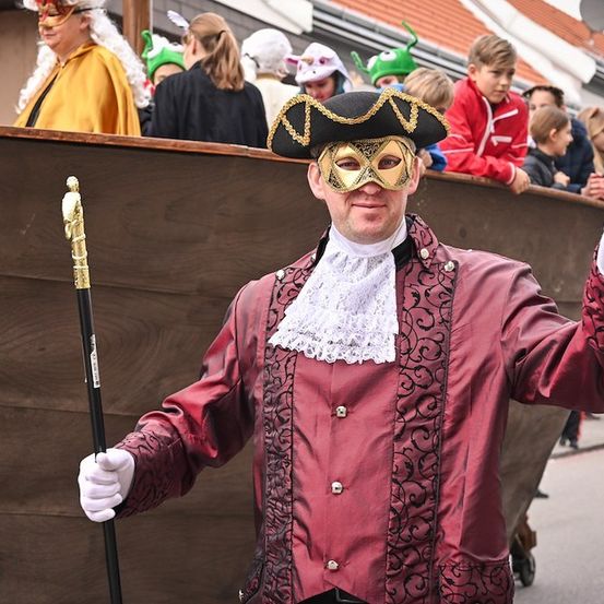 Ein Mann in einem roten Kostüm und einer Maske winkt in die Kamera, während er einen Stock hält. Hinter ihm ist ein hölzernes Boot zu sehen, auf dem Menschen in Kostümen zusehen.