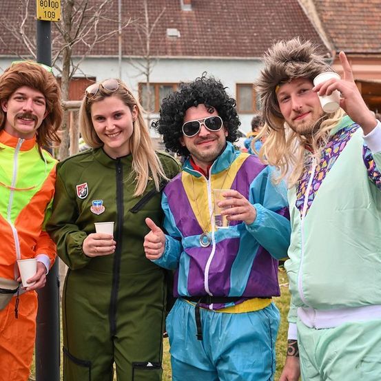 Vier Personen in farbenfrohen 70er-Jahre-Outfits stehen auf einer Wiese, lächeln und halten Tassen. Hinter ihnen ist ein Gebäude mit einem braunen Dach und einem Schild mit der Aufschrift 80109 zu sehen.