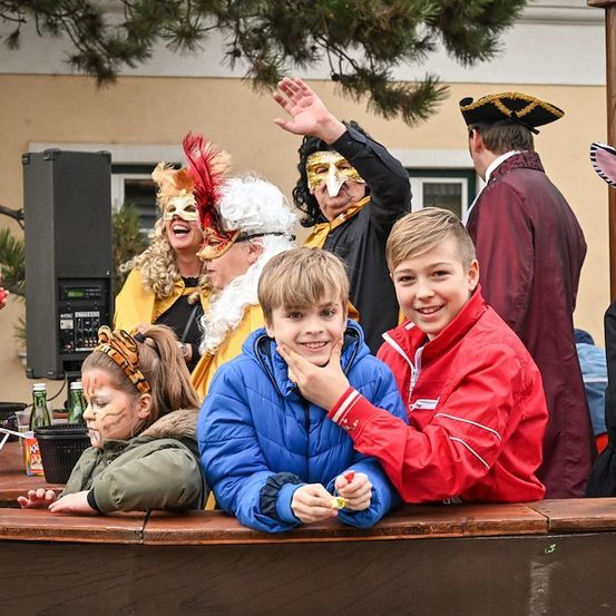 Eine Gruppe von Kindern und Erwachsenen in Kostümen versammelt sich um einen Tisch, möglicherweise für eine festliche Veranstaltung. Einige lächeln und posieren für ein Foto. Dahinter befindet sich ein Gebäude mit einem Fenster und einem Baum.