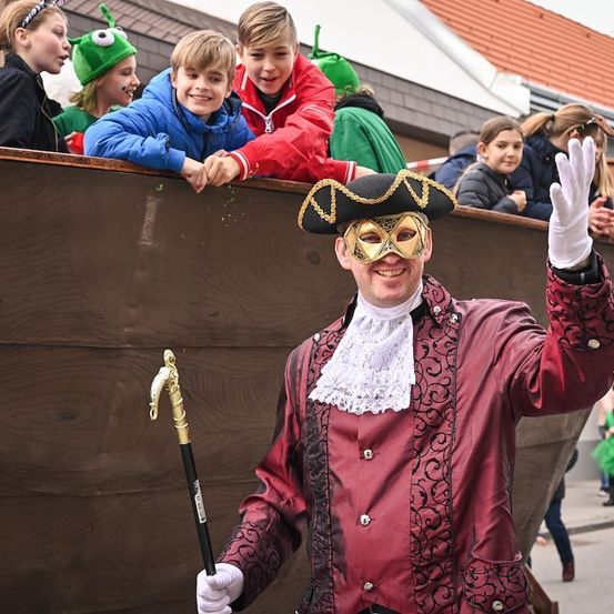 Ein Mann in einem roten Kostüm und einer Maske winkt zur Kamera, während er einen Stock hält. Hinter ihm sehen Kinder in Kostümen von einem Boot aus zu.