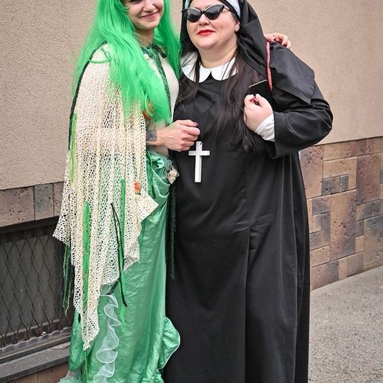 Zwei Frauen in Kostümen, eine in einem grünen Meerjungfrauen-ähnlichen Outfit und die andere in einer Nonnentracht, stehen eng beieinander auf einem Bürgersteig.