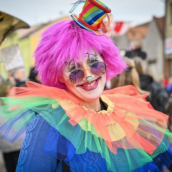 Eine Person in einem farbenfrohen Clownkostüm mit rosa Haaren und buntem Make-up steht auf einer Straße, umgeben von einer Menge.