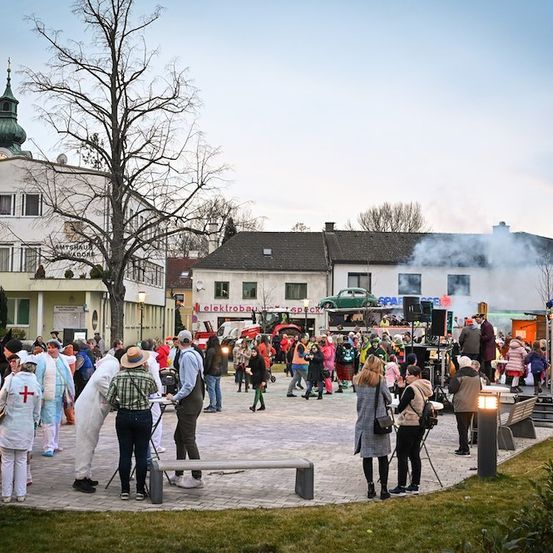 Ein lebhaftes Ereignis auf einem Stadtplatz mit Menschen in Kostümen, einem Feuerwehrwagen und einer Bühne mit Rauch.