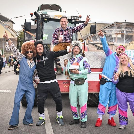 Eine Gruppe von Menschen in bunten Kostümen posiert für ein Foto vor einem Traktor auf einer Straße.