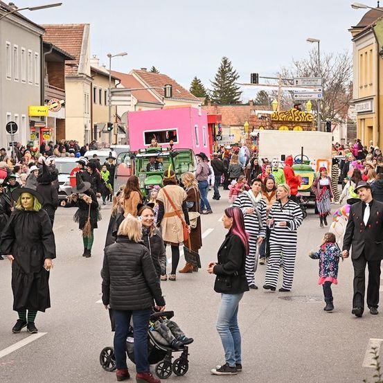 Eine Straße voller Menschen in Kostümen, einige gehen, andere stehen, mit einem Umzug im Hintergrund, geparkte Autos am Straßenrand und Straßenlaternen und Gebäude in der Ferne.