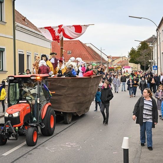 Ein Umzug mit einem Wagen, auf dem Leute in Kostümen sitzen, gezogen von einem roten Traktor, auf einer Stadtstraße mit Zuschauern.